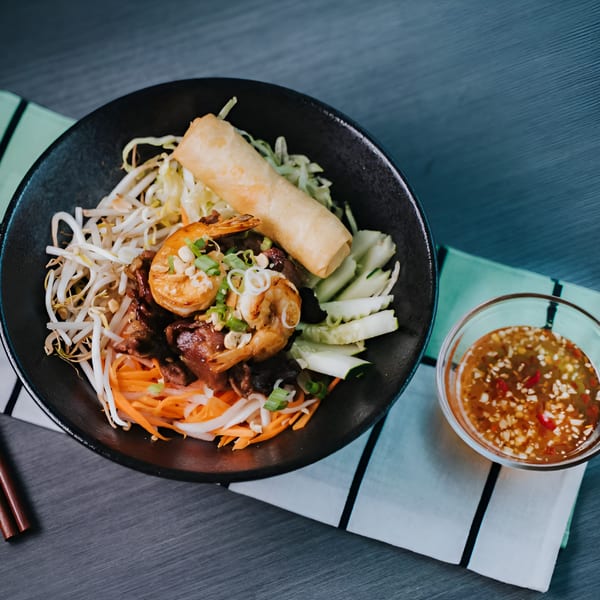 Vietnamese vermicelli bowl with shrimp, beef, spring roll, and vegetables, with a side of dipping sauce.