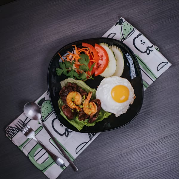 Vietnamese dish with beef, shrimp, fried egg, and fresh vegetables on a black plate.