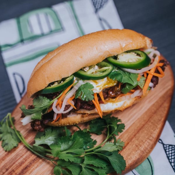 A Vietnamese Banh Mi sandwich with meat, egg, jalapeños, cilantro, and pickled vegetables on a wooden board.
